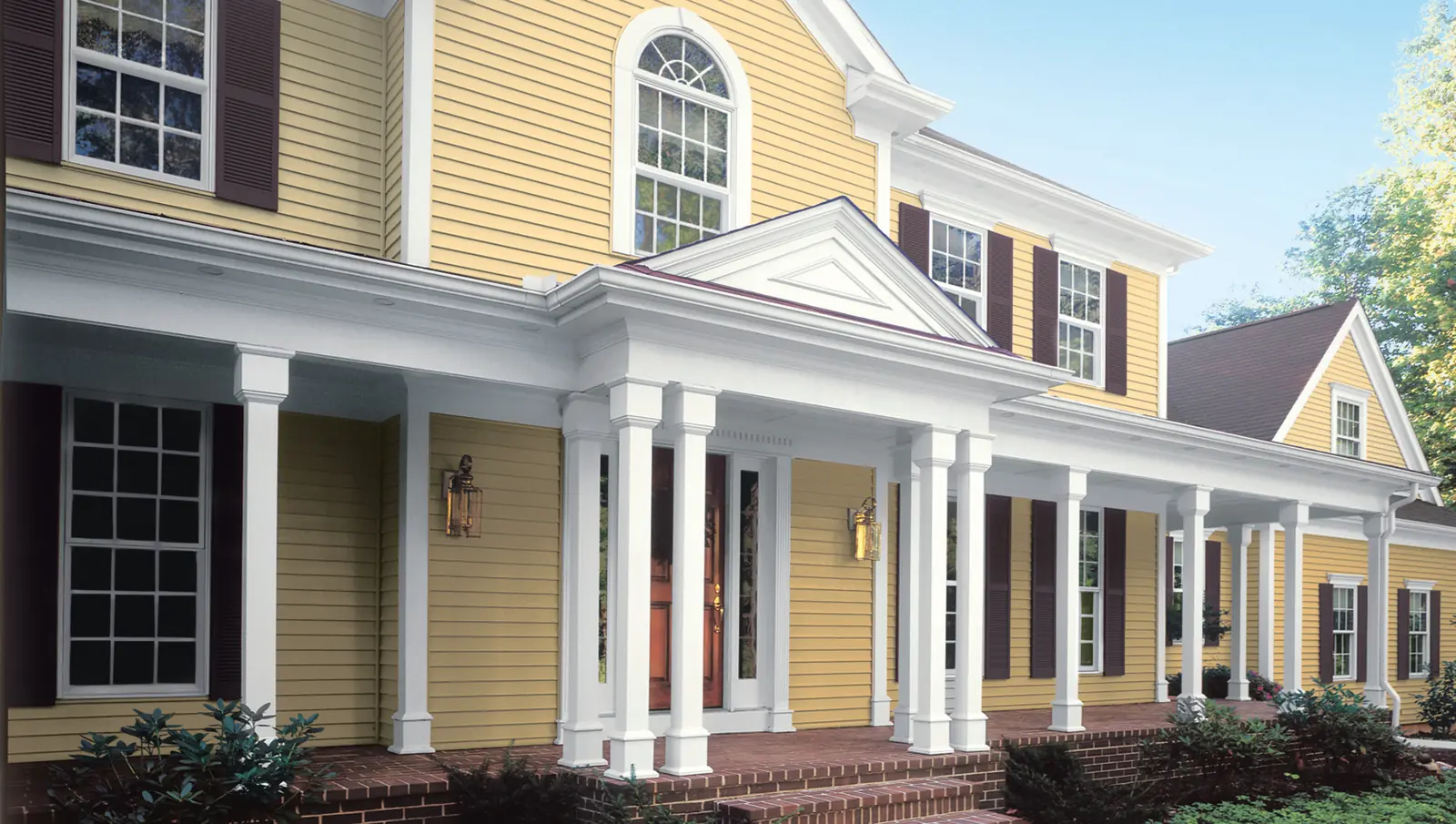 Yellow two-story colonial with Mastic vinyl lap siding, dark shutters, and a long white-columned porch